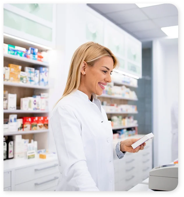 Uśmiechnięty farmaceuta w białym fartuchu trzyma pudełko z lekami przy nowoczesnej ladzie aptecznej. W tle widoczne są półki z produktami i inny farmaceuta.